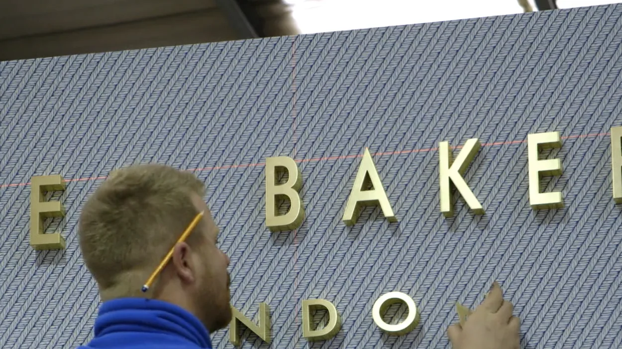 An installer fixing brass Ted Baker letters to a patterned wall panel, with a pencil behind his ear.